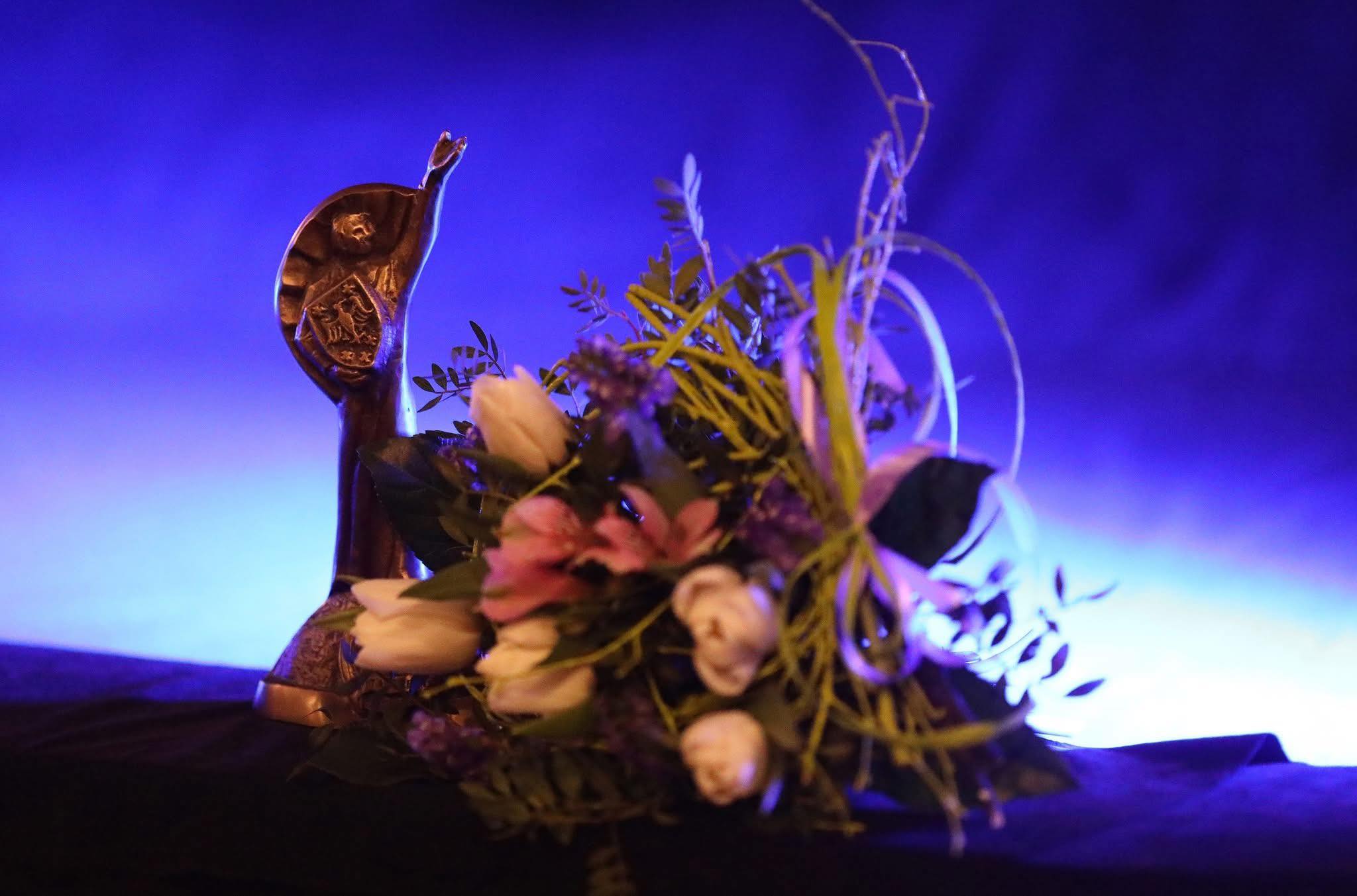 Na niebieskim tle statuetka metalowego  Clemensa i bukiet kwiatów