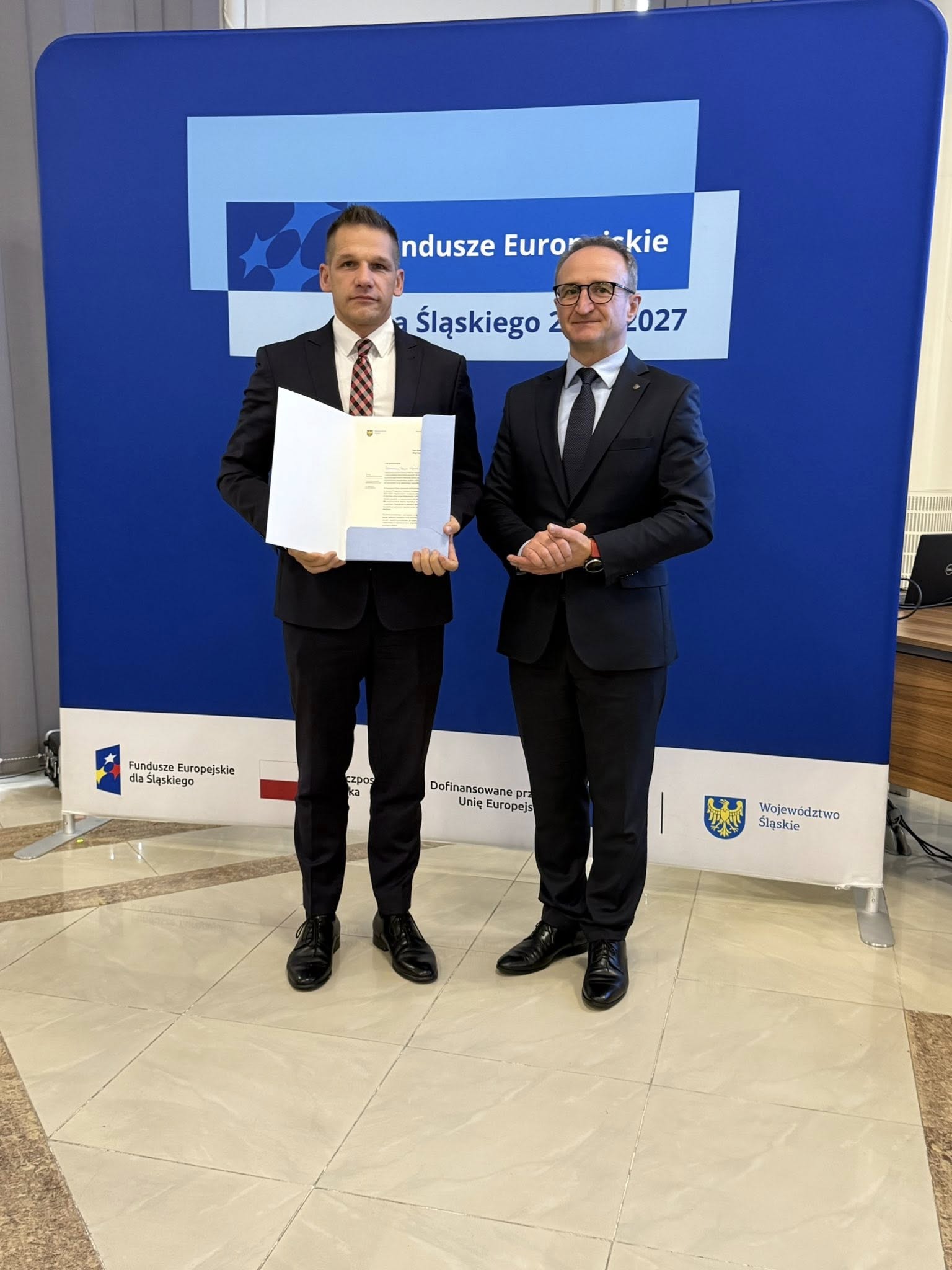 Dwóch mężczyzn w pomiesczzeniu pozujących do zdjęcia. Jeden z nich trzyma teczkę z listem gratulacyjny. 