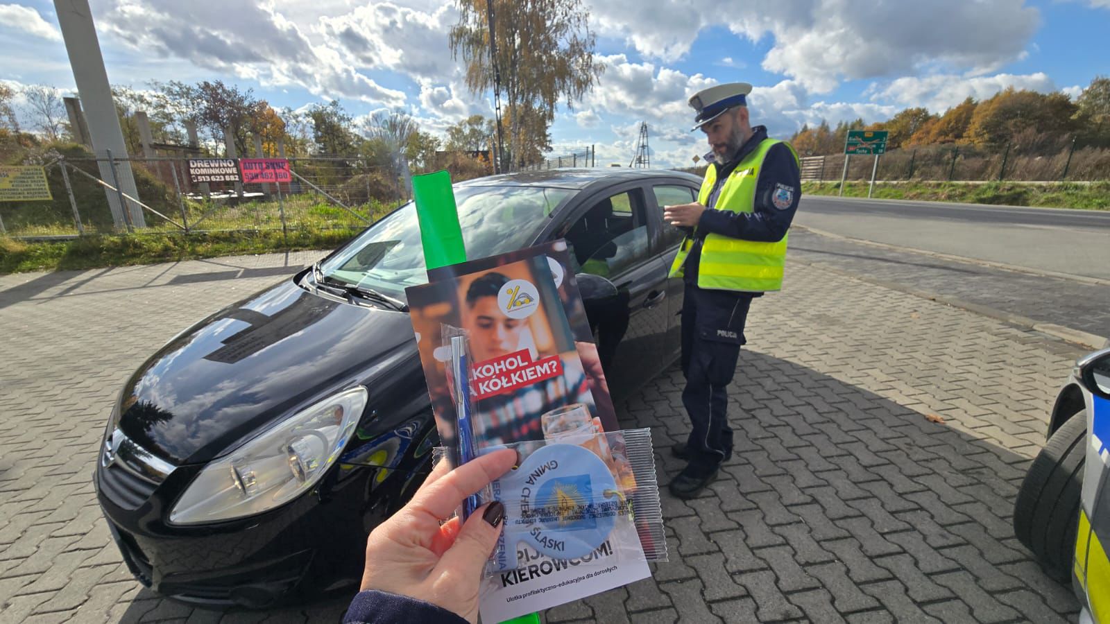 Na zdjęciu policjant kontolujacy samochód/ w dłoni materiały edukacyjne