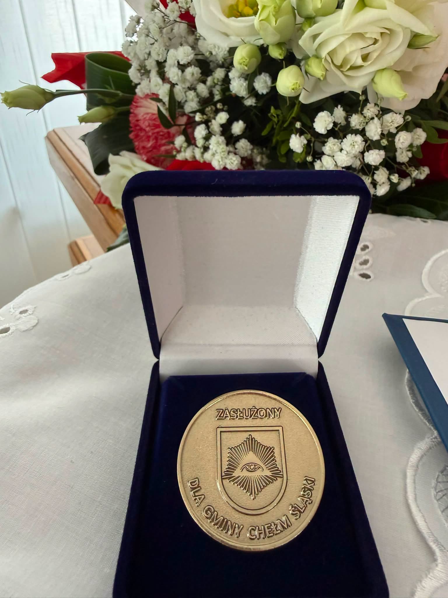 Medal w opakowaniu leżacy na stoliku. Za nim bukiet kwiatów. 