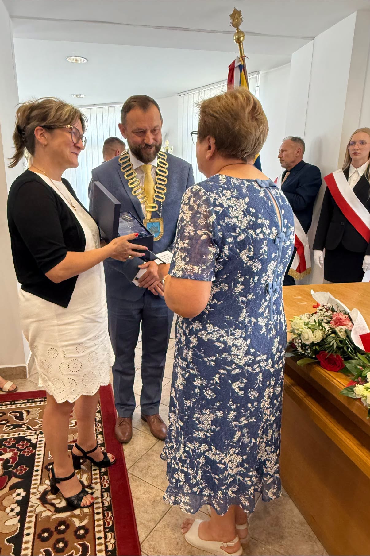 Mężczyzna i kobieta składający życzenia kobiecie w pomieszczeniu. Wręczają statuetkę. 