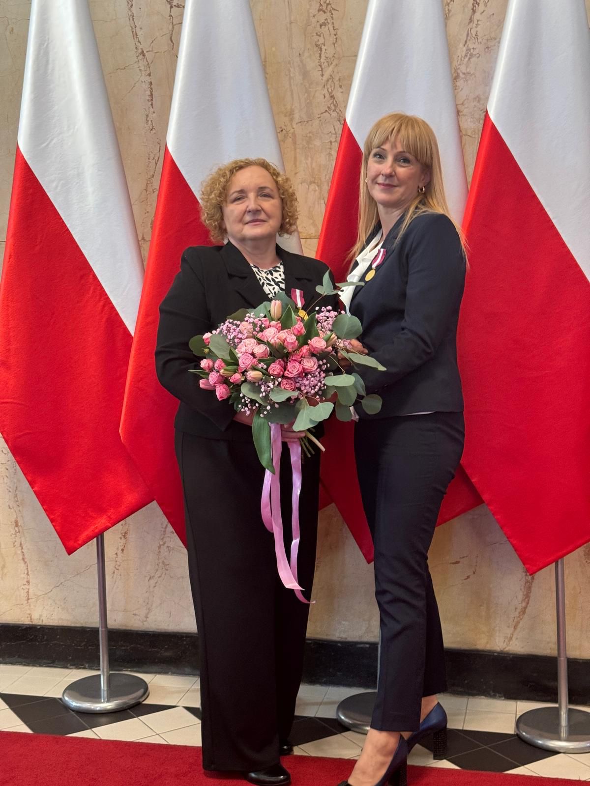 Dwie kobiety pozujące do zdjęcia na tle flag Państwowych z bukietem kwiatów 