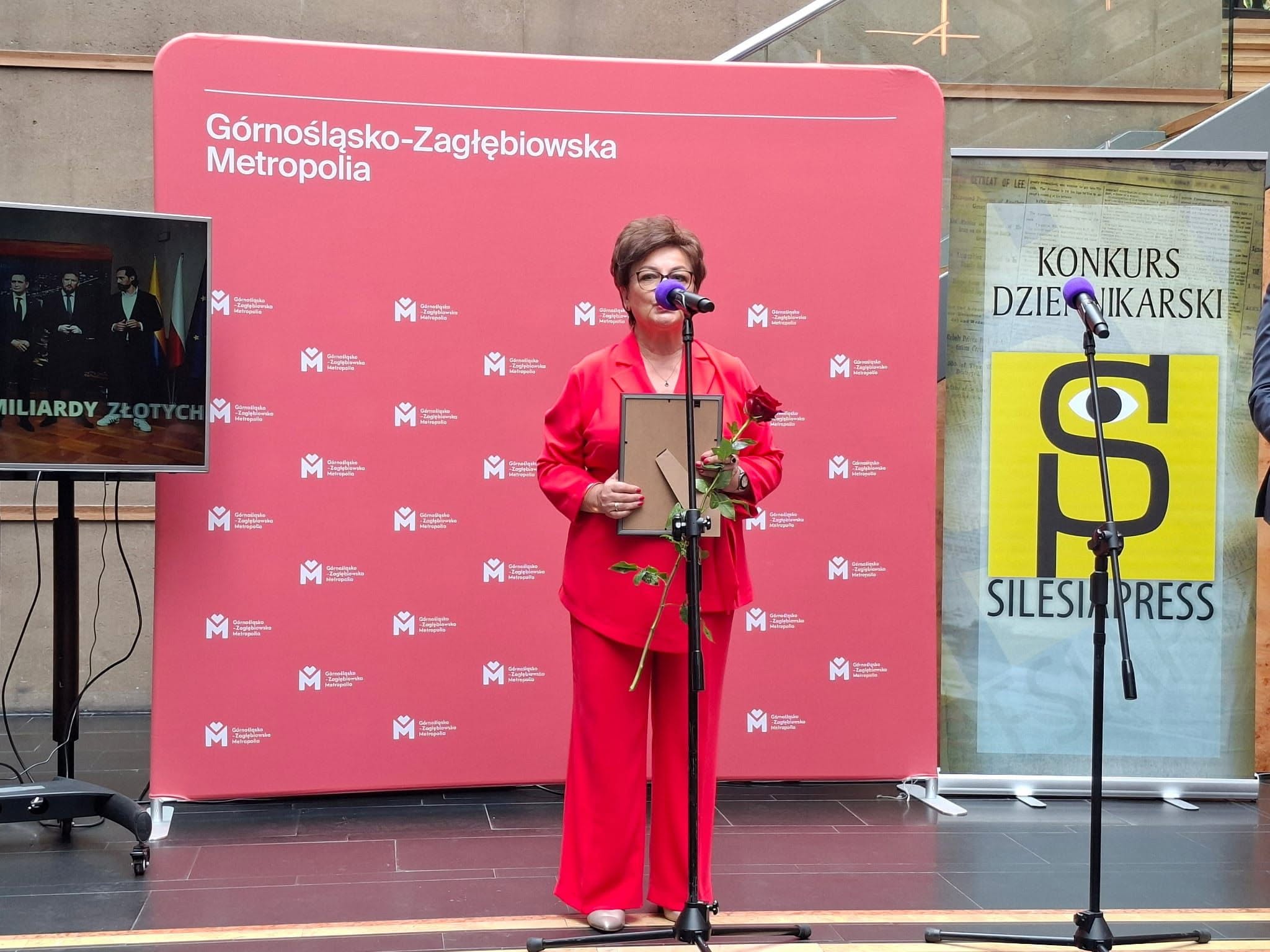 Kobieta trzymająca w dłoni obraz, różę i przemawiająca przed ścianką reklamową. 