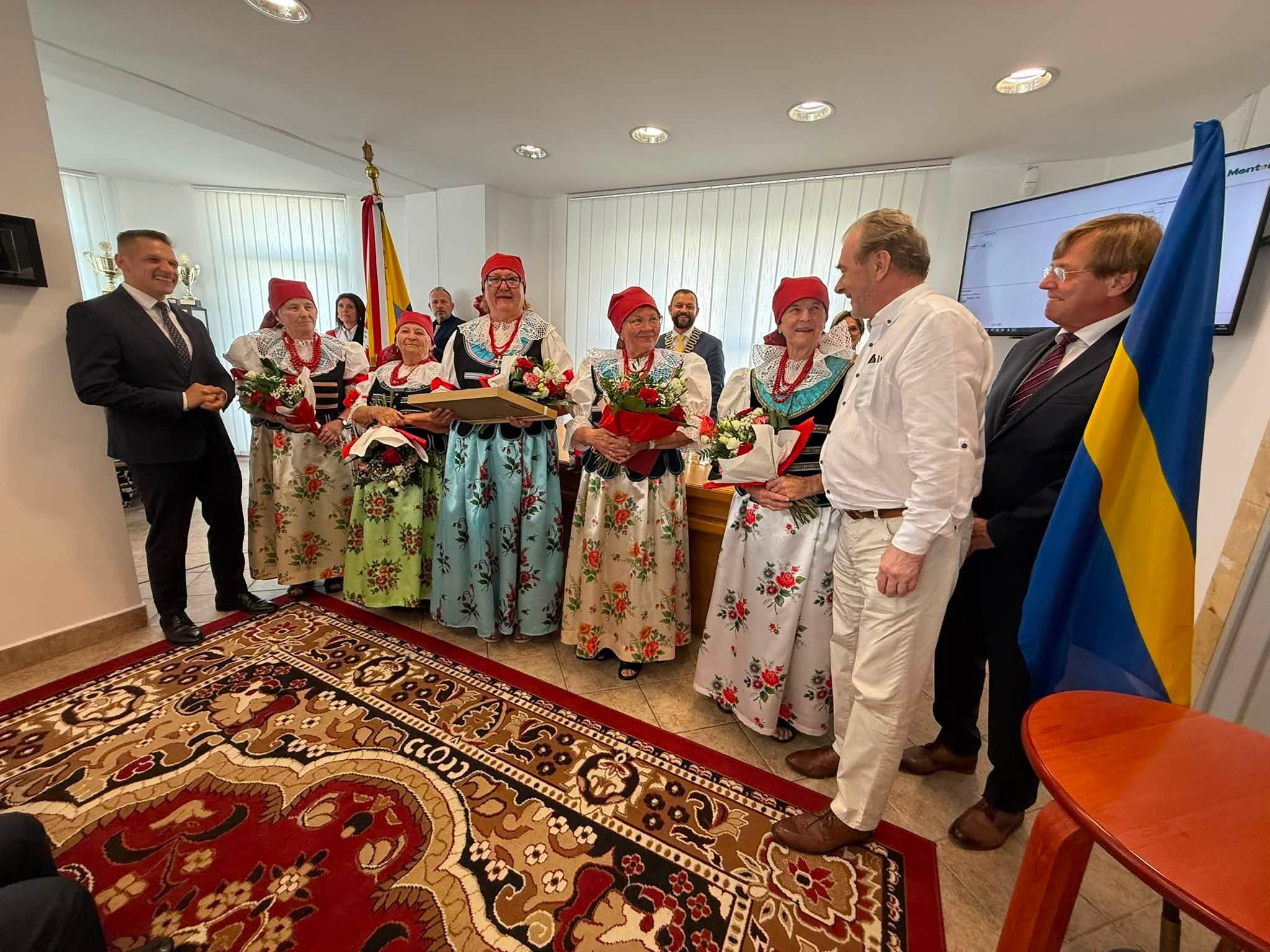 Osoby pozujące do zdjęcia w tym panie w strojach ludowych. Wszyscy są w pomieszczeniu. 