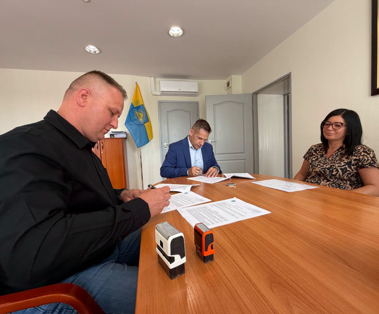 Dwaj mężczyźni w pomieszczeniu podpisujący umowę i przyglądająca się im kobieta. 