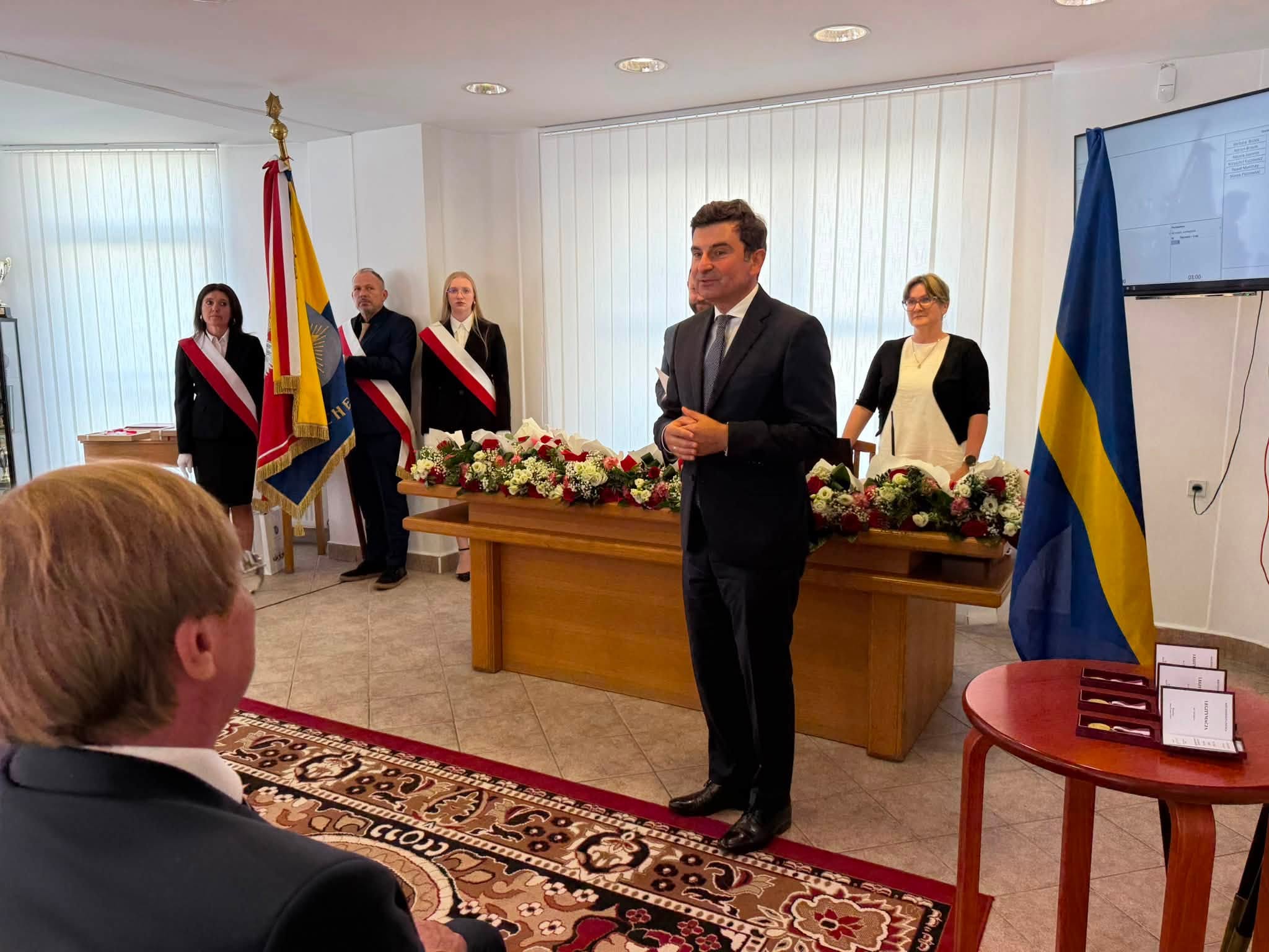 Mężczyna przemawiający w pomieszczeniu. W tle poczet sztandarowy i stojąca kobieta