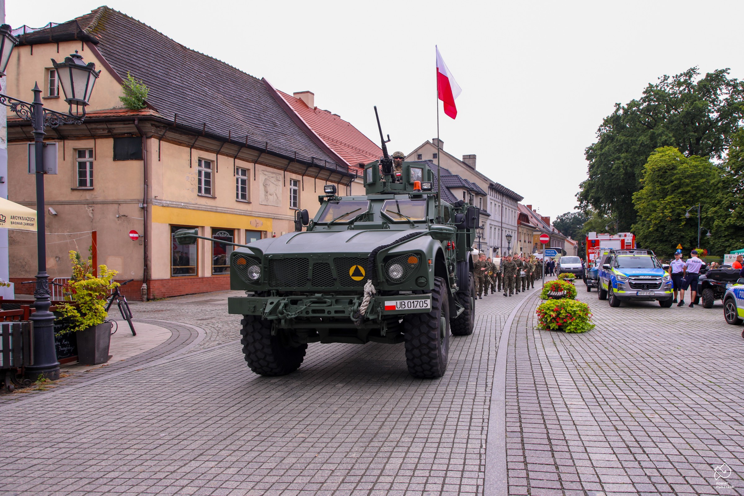 Wóz wojskowy na rynku a za nim przemarsz uczestników. 