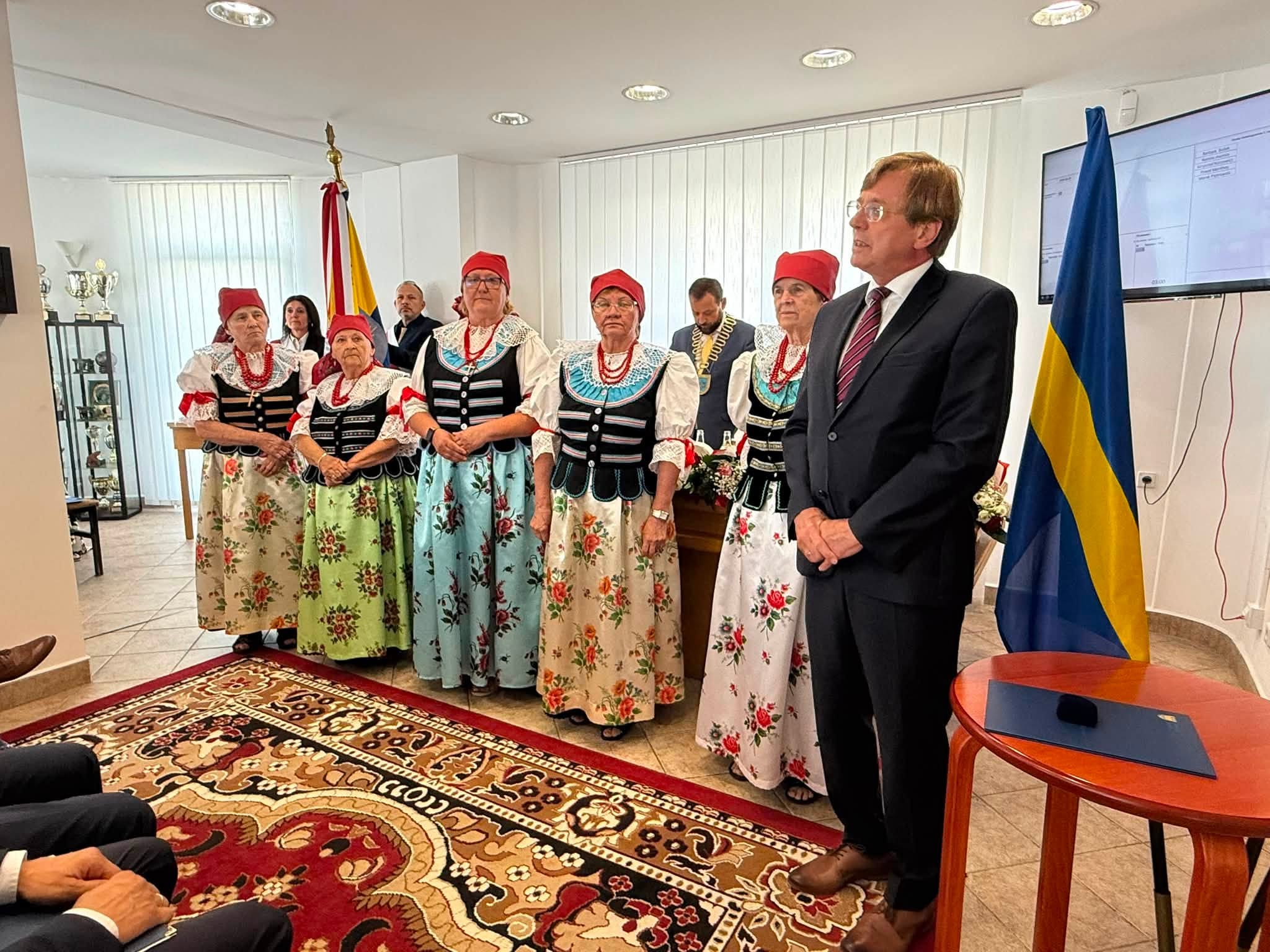 Kobiety w strojach ludowych stojące w pomieszczeniu i mężczyzna przemawiający do zgromadzonych uczestników spotkania. 