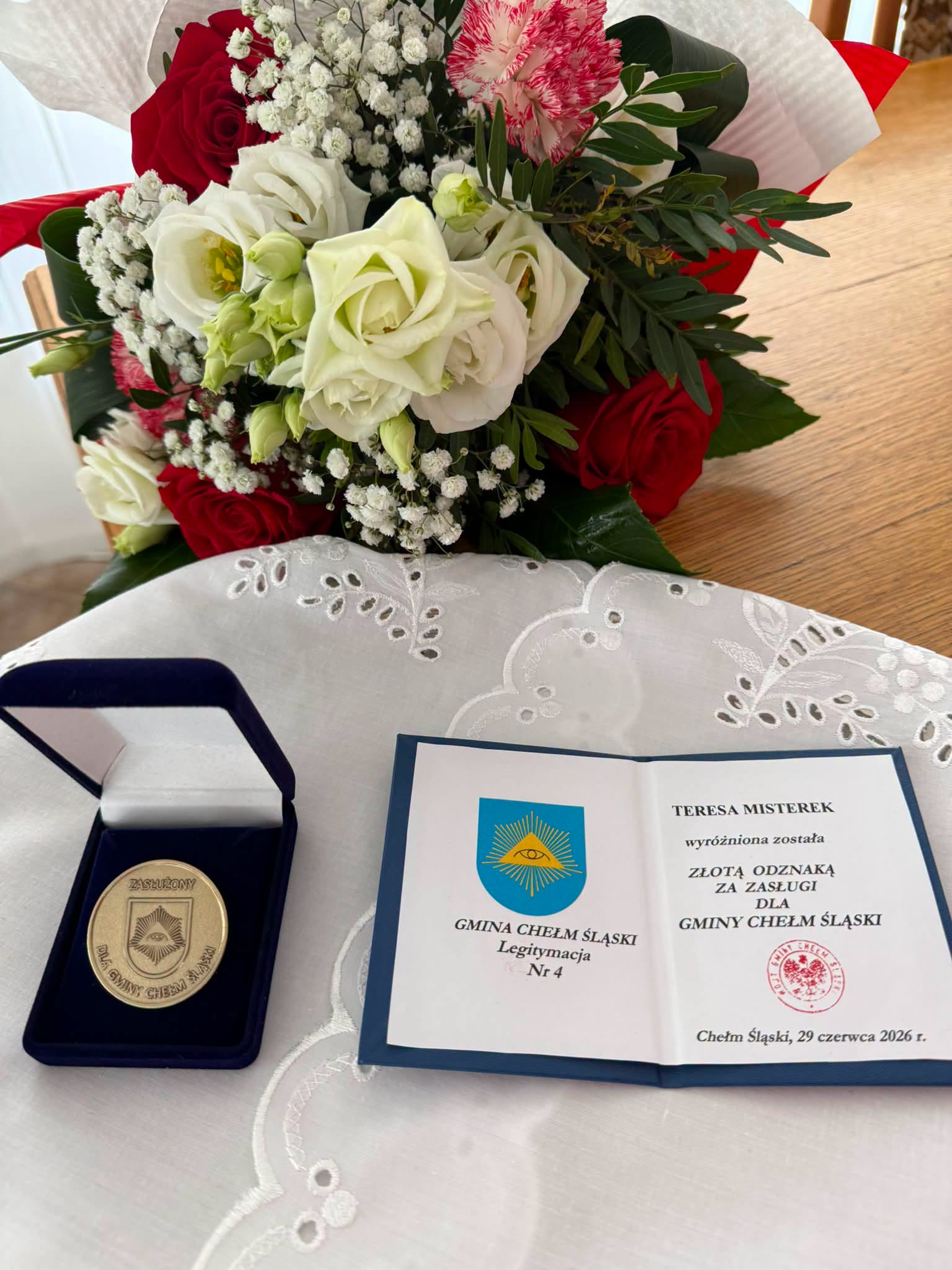 Legitymacja i medal leżace na stole a za nimi bukiet kwiatów. 