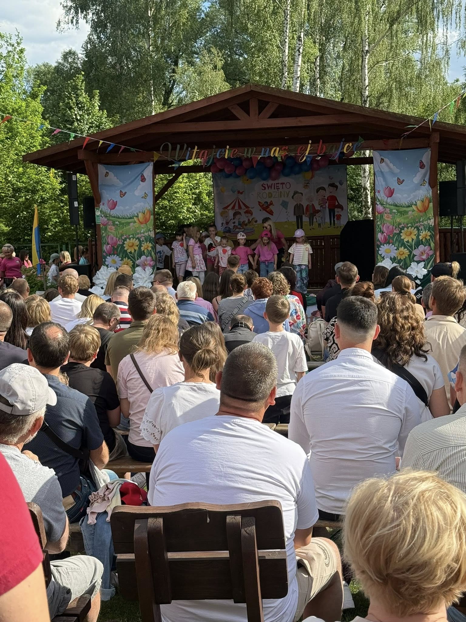 Osoby siedzące przed sceną a na scenie występujące dzieci przedszkolne. 