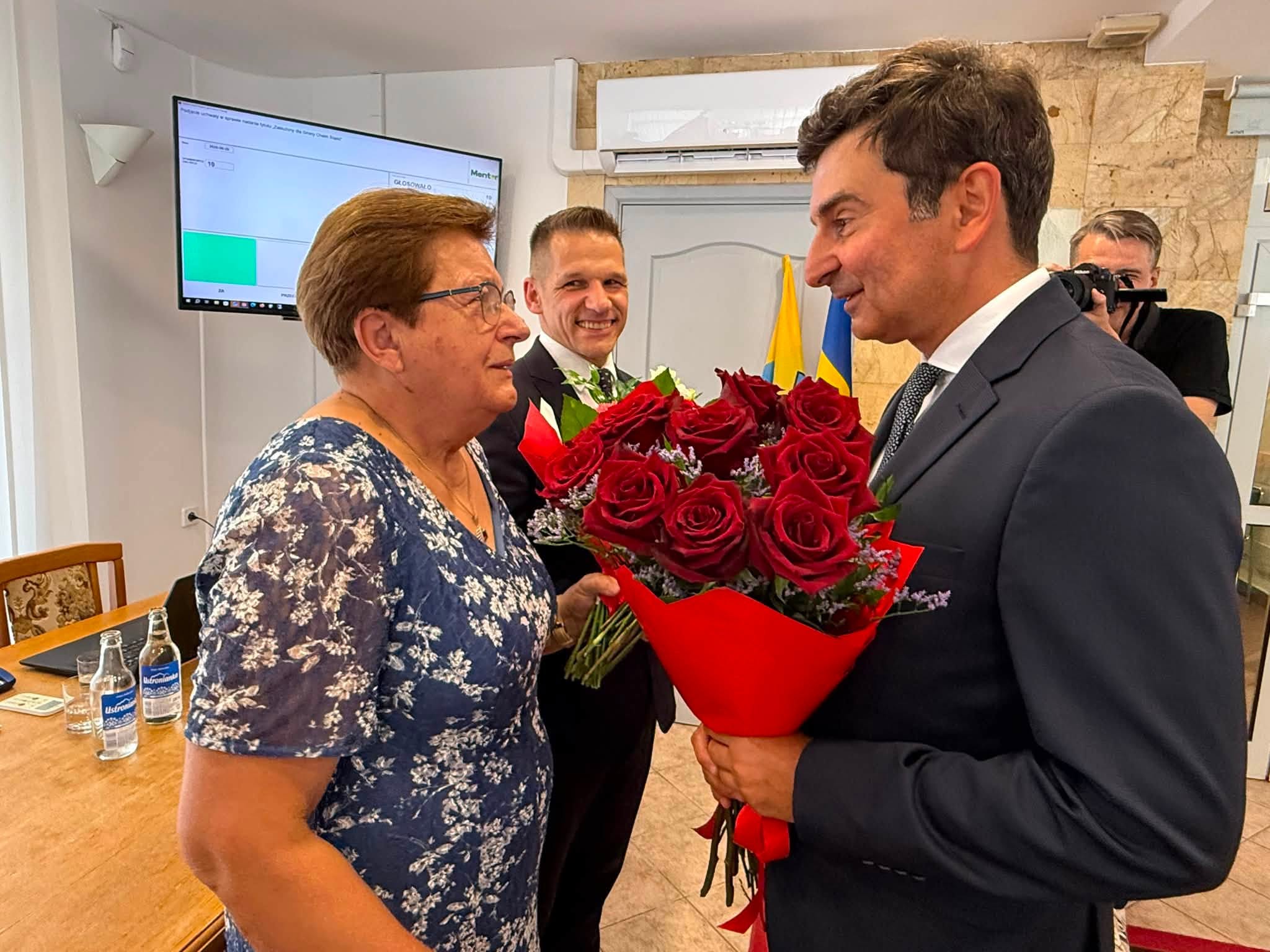 Mężczyzna wręczający wiązankę kwiatów kobiecie. W tle dwaj mężczyżni. 