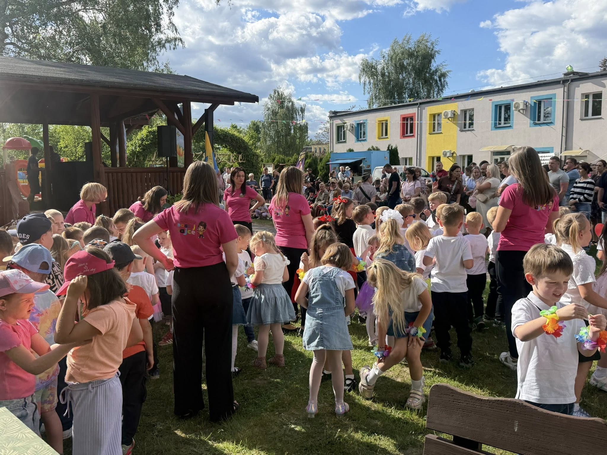 Dzieci oczekujące przed sceną na swój występ wraz z opikunkami. W tle osoby starsze siedzące jako widownia